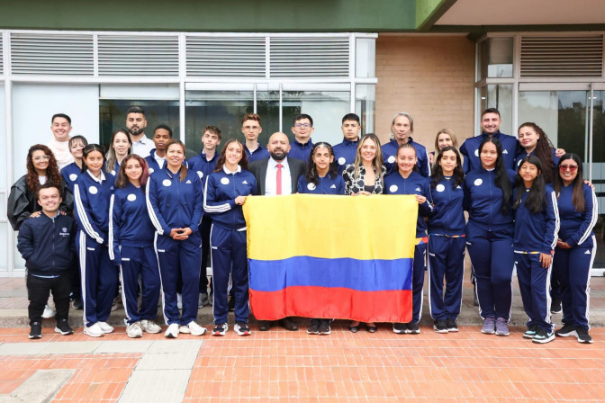 Grupo de deportistas colombianos en sudaderas azules sostienen la bandera de Colombia junto a funcionarios institucionales.
