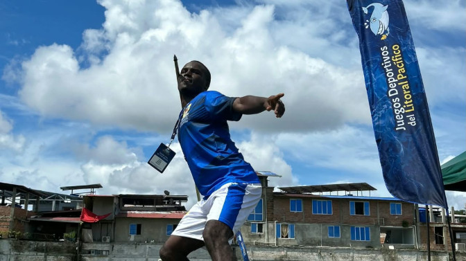 Atleta masculino en posición de lanzamiento de jabalina durante los Juegos Deportivos del Litoral Pacífico, con bandera oficial del evento al fondo.